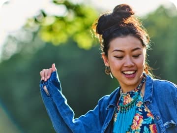 A woman dancing outside, smiling and wearing a denim jacket