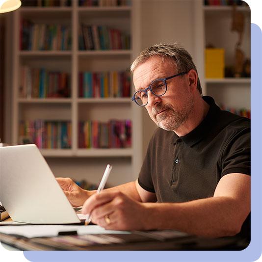 Man sitting by a desk at home writing whilst using a laptop