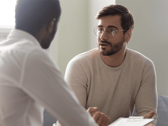 man with glasses talking to another man, both sitting down