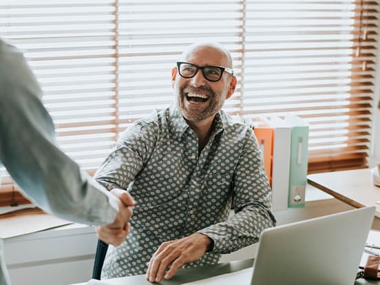 Bald man in glasses smiles while shaking hands across a desk with a laptop, in a sunlit office with wooden blinds.