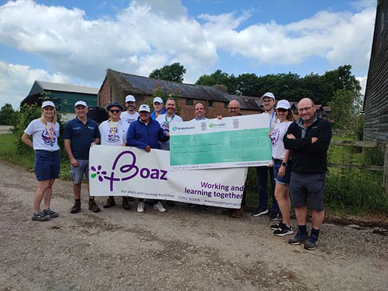 Group of people holding a large check and a "Boaz" banner outdoors, with a barn and cloudy sky in the background.