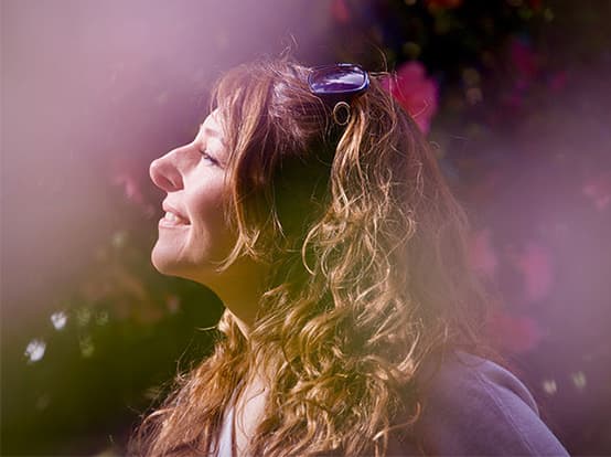 Woman with curly hair and sunglasses on head, smiling while looking up, surrounded by a dreamy pink and blurred floral background.