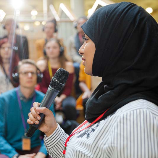A woman wearing a hijab, speaking into a microphone to audience